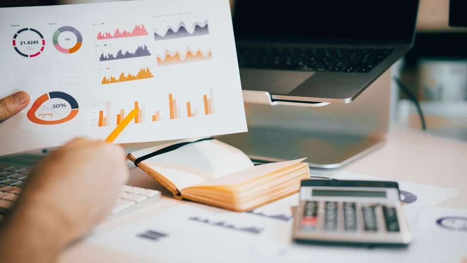 A hand pointing a pencil at charts on a printed report, with a calculator and laptop