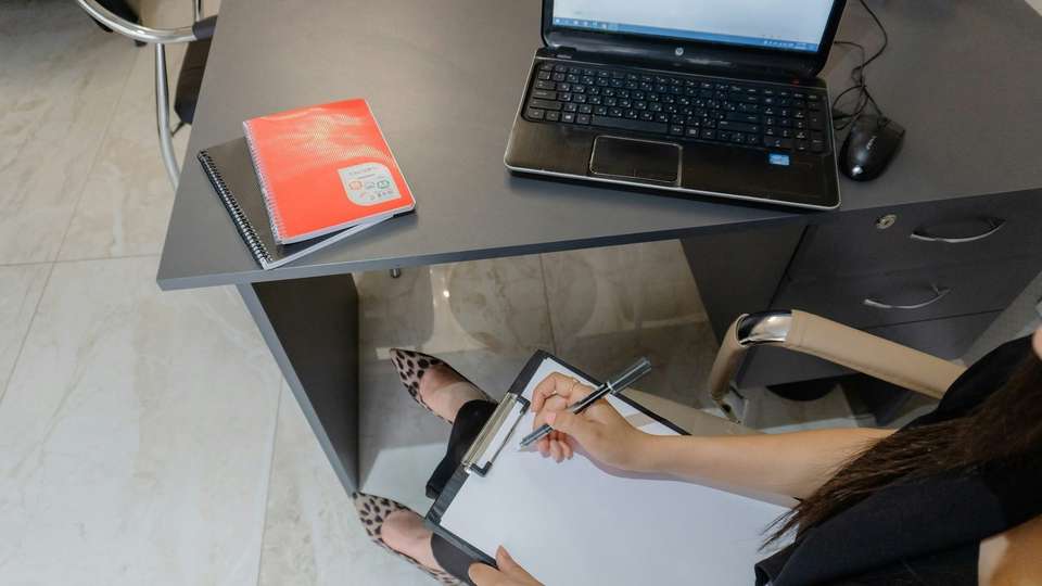 A person filling in a form on a clipboard beside a desk with a laptop