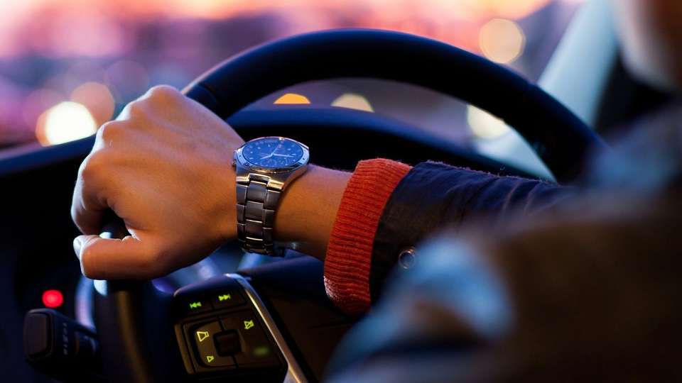 A driver's hand on the steering wheel at dusk, with city lights ahead