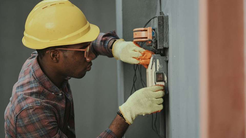An electrician in a yellow hard hat and gloves installing wiring on a wall