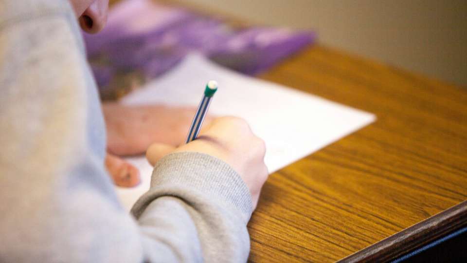 A student writing with a pencil on a sheet of paper at a school desk