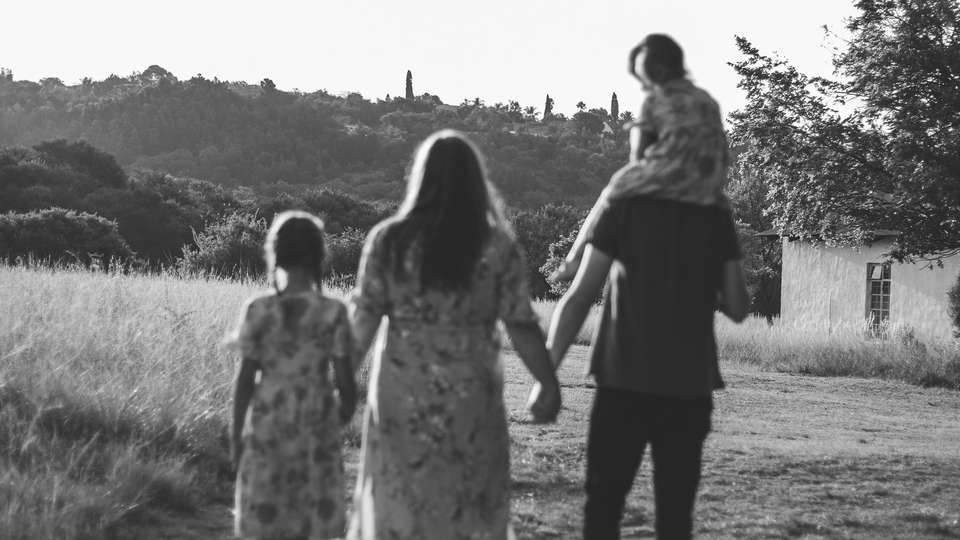 A family of four walking hand in hand across a grassy field, seen from behind, in black and white