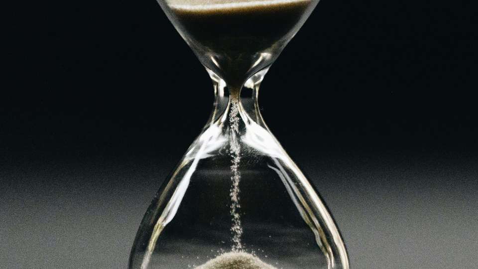 Sand running through the neck of a glass hourglass against a dark background