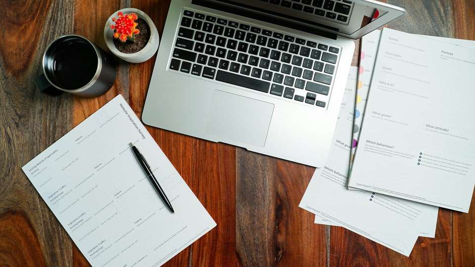 A laptop, printed pages and a pen on a wooden desk, seen from above