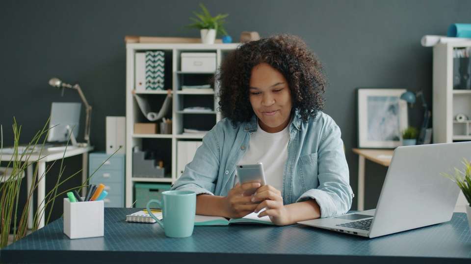 A young woman smiling at her phone at a desk, with a laptop, a mug and an open notebook beside her