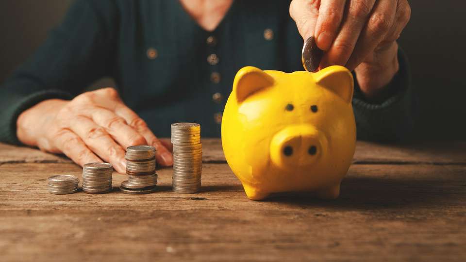 An older person's hand dropping a coin into a yellow piggy bank, next to small stacks of coins on a wooden table