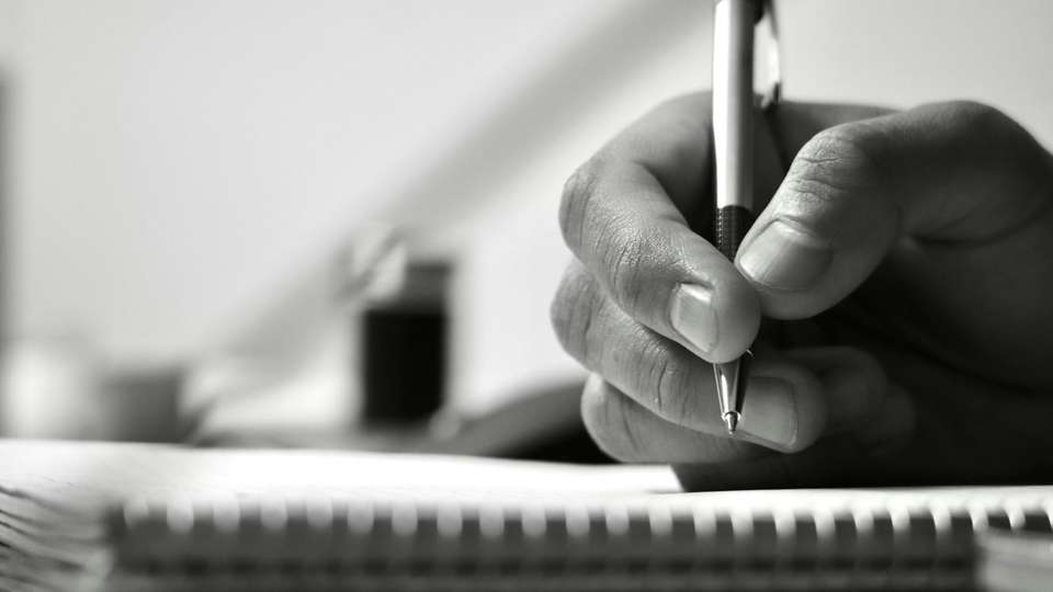 A hand writing in a notebook with a pen, in black and white