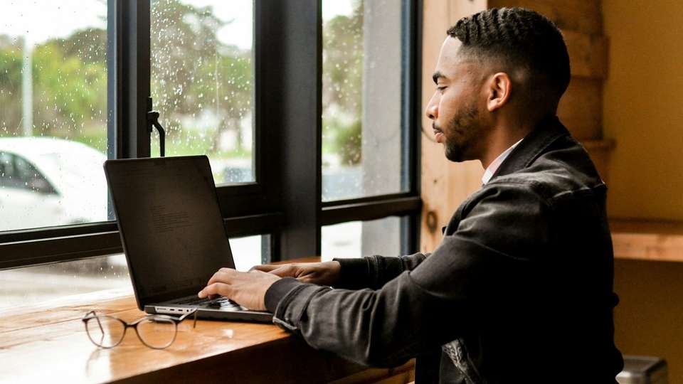 A man typing on a laptop at a wooden counter by a window